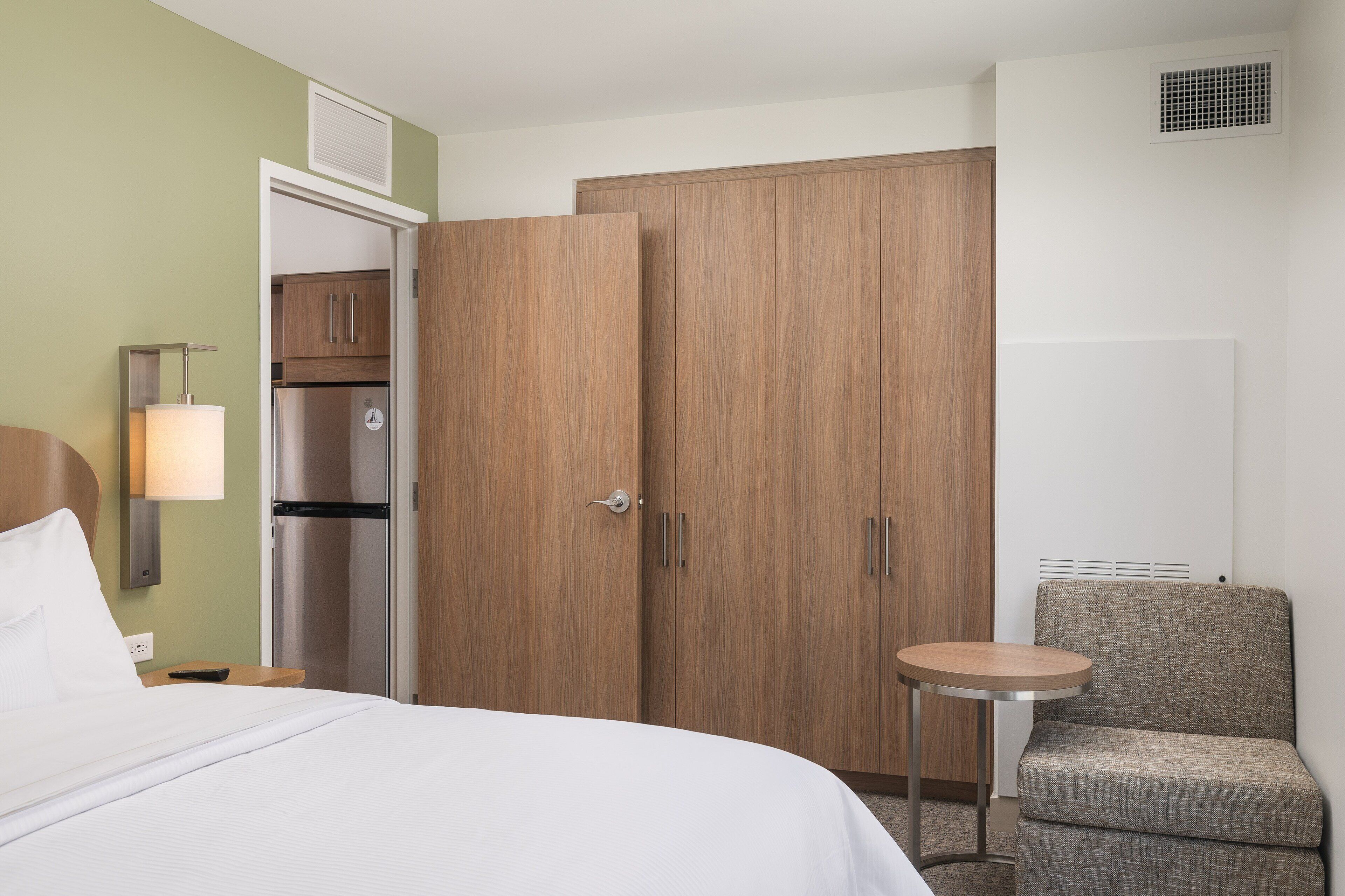 A hotel room with a bed, a small round table, a chair, a wardrobe, and a refrigerator.
