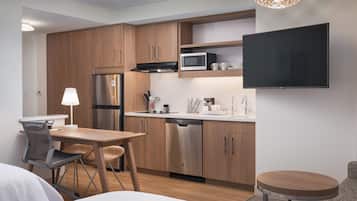 A compact kitchen with wooden cabinets, a stainless steel refrigerator, a microwave, and a dishwasher. There's a dining table with chairs, a small round table, and a TV mounted on the wall.