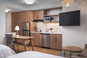 A compact kitchen with wooden cabinets, a stainless steel refrigerator, a microwave, and a dishwasher. There's a dining table with chairs, a small round table, and a TV mounted on the wall.