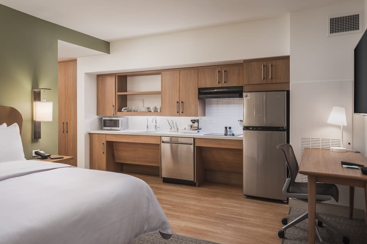 A hotel room with a bed, a desk, a chair, a kitchenette with a refrigerator, microwave, and sink, and a lamp.