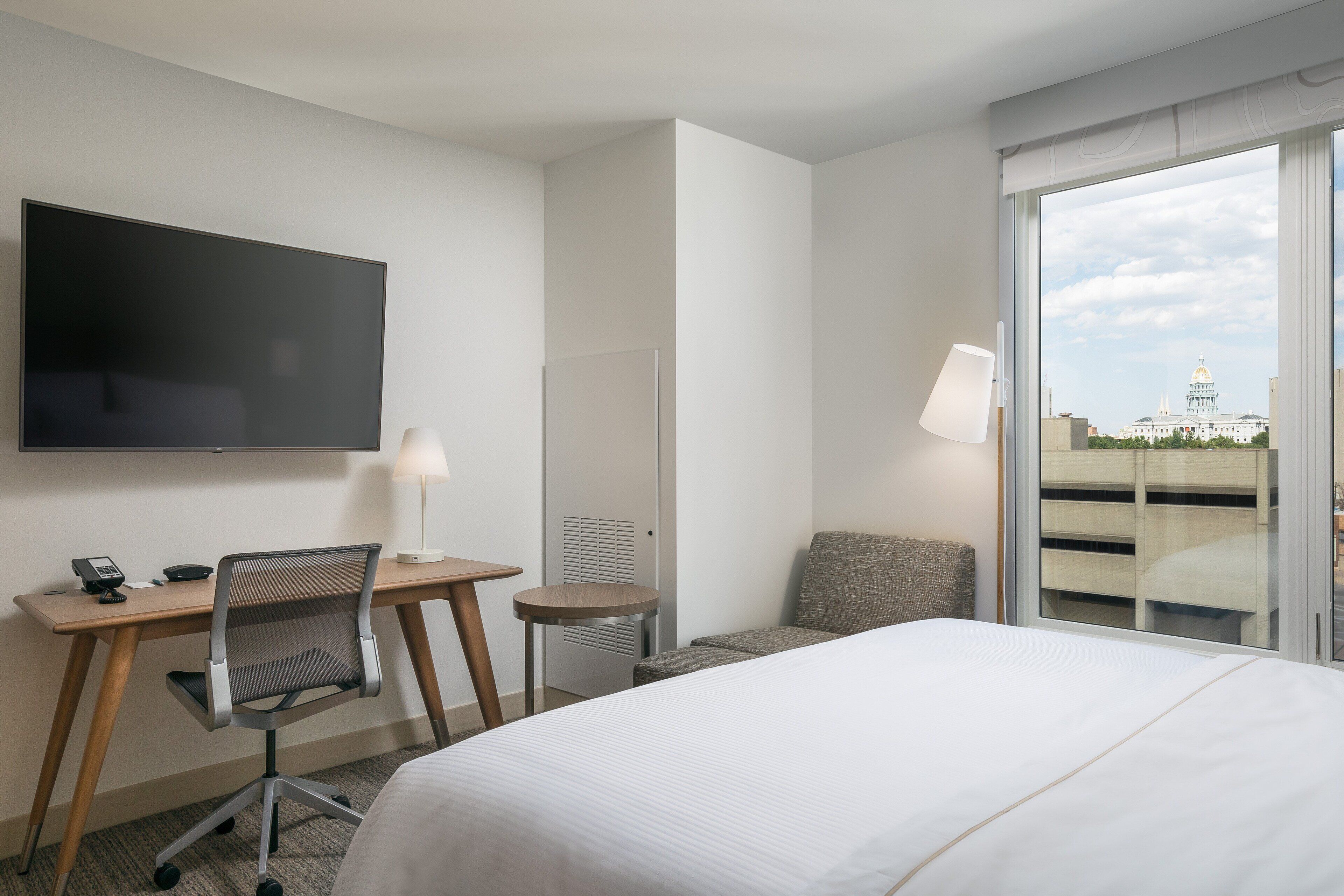 Ein Hotelzimmer mit Bett, Schreibtisch mit Stuhl, Fernseher und Blick auf ein Gebäude mit einer Kuppel durch ein großes Fenster.