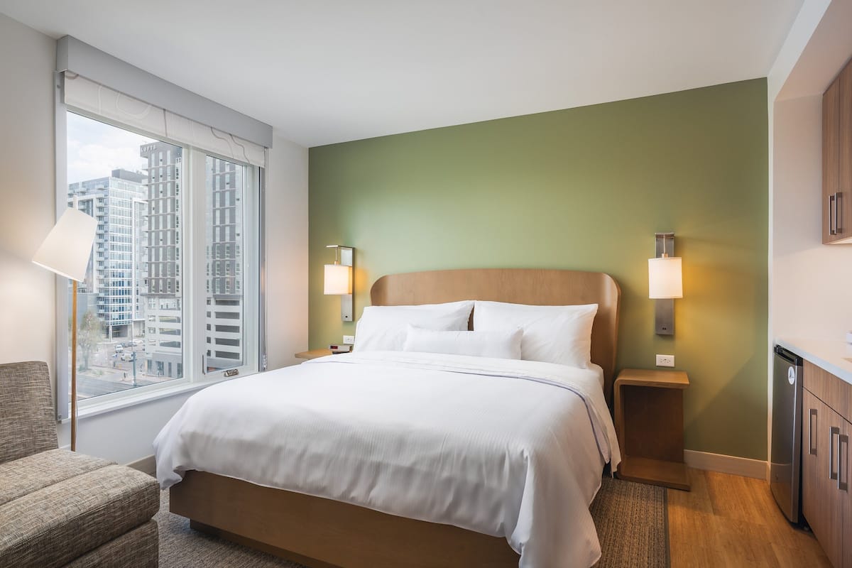 A hotel room with a large bed, two bedside tables with lamps, a view of buildings through a window, and a small seating area.