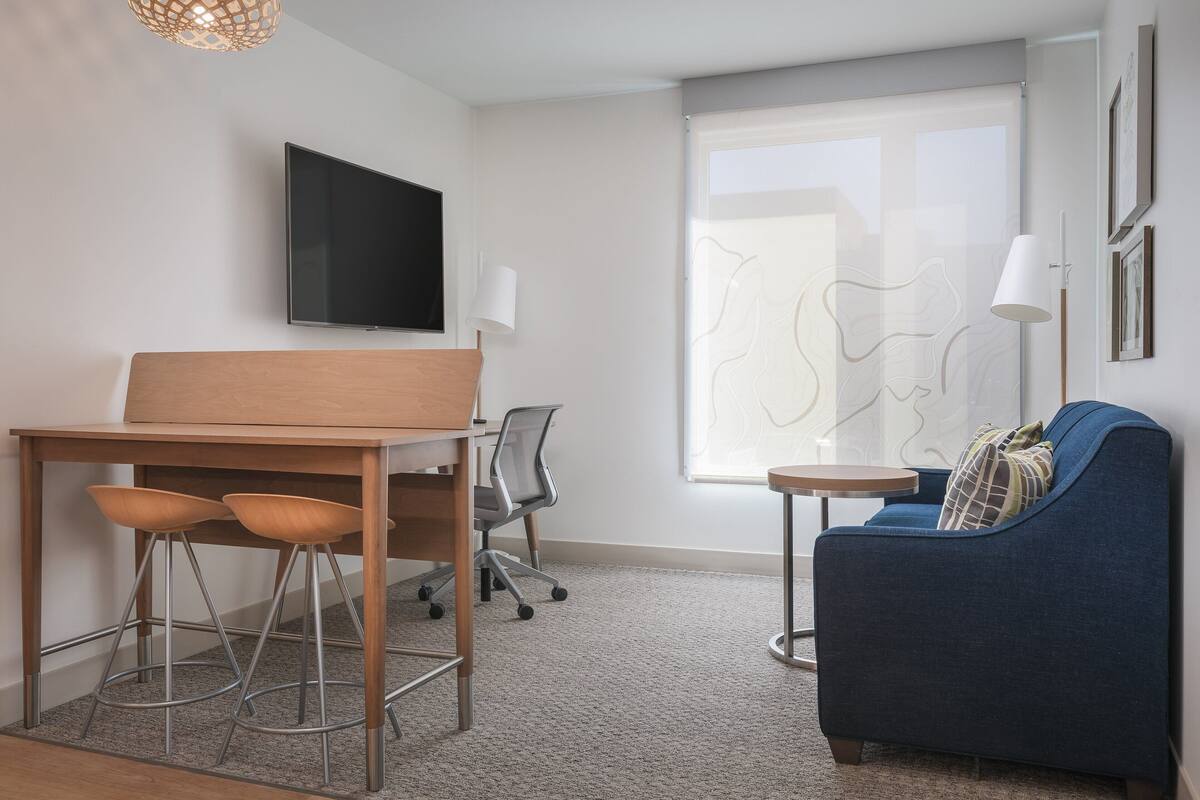 A modern room with a wooden desk, two bar stools, a blue sofa, a small round table, a TV mounted on the wall, and a large window with a view of the outside.