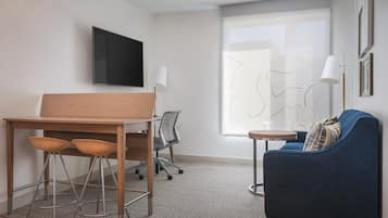 Ein modernes Zimmer mit Schreibtisch aus Holz, zwei Barhocker, einem blauen Sofa, einem kleinen runden Tisch, einem an der Wand montierten Fernseher und einem großen Fenster mit Außenblick.