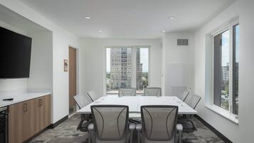 Un salón de conferencias con una mesa grande, sillas, un televisor y vistas a edificios a través de una ventana.