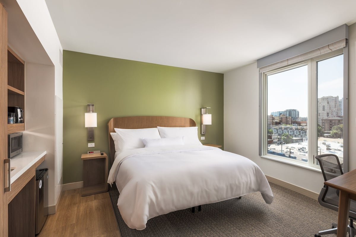 A hotel room with a bed, bedside tables, a desk, and a large window offering a city view.