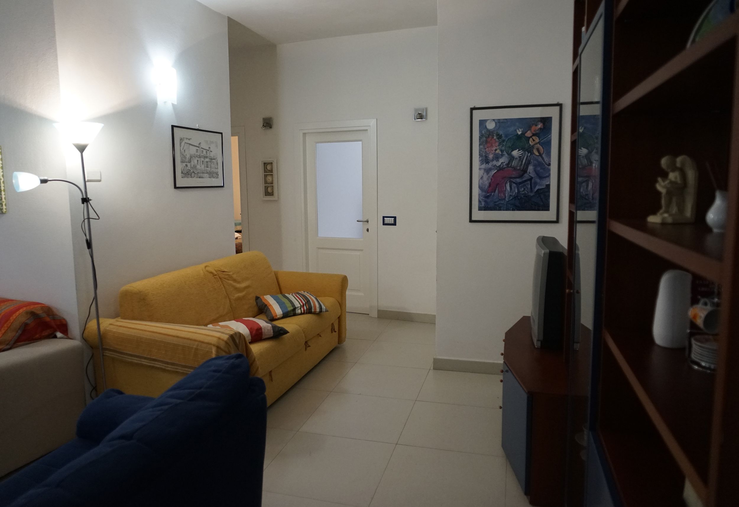 Uma sala de estar com um sofá amarelo, um sofá azul, um abajur e uma estante com objetos decorativos.