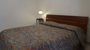 A single bed with a patterned bedspread and a wooden headboard. A small bedside table with a lamp is visible to the left of the bed. - Casa Granchio (Alghero)