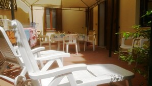 Een terras met witte plastic stoelen en een tafel, een raam met houten luiken, en een plant aan de rechterkant.