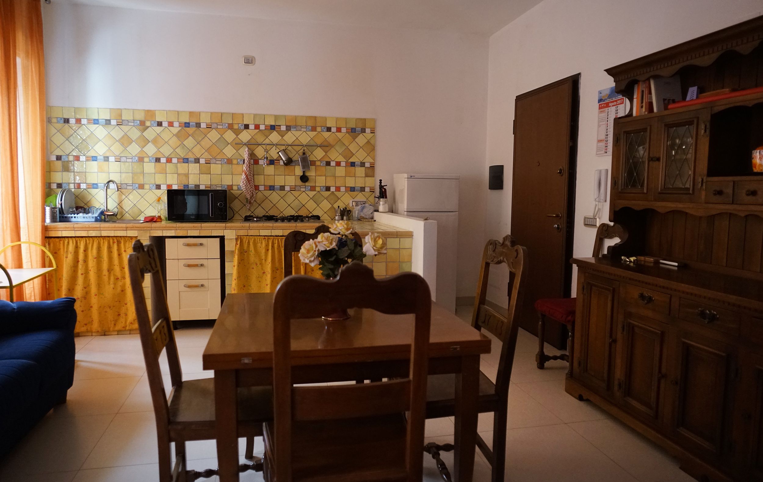 A compact kitchen with a tiled backsplash, a microwave, a stove, and a refrigerator. There is a dining table with chairs and a wooden cabinet with glass doors.