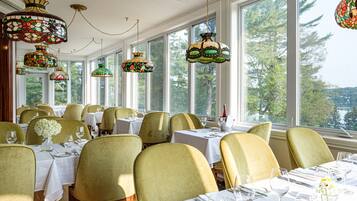 A restaurant with multiple tables set for dining, large windows with a view of trees and a lake, and colorful stained glass lamps.