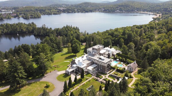 A large, multi-story building with a swimming pool, surrounded by lush greenery and a lake. - StoneHaven Le Manoir Relais & Châteaux (Sainte-Agathe-des-Monts)