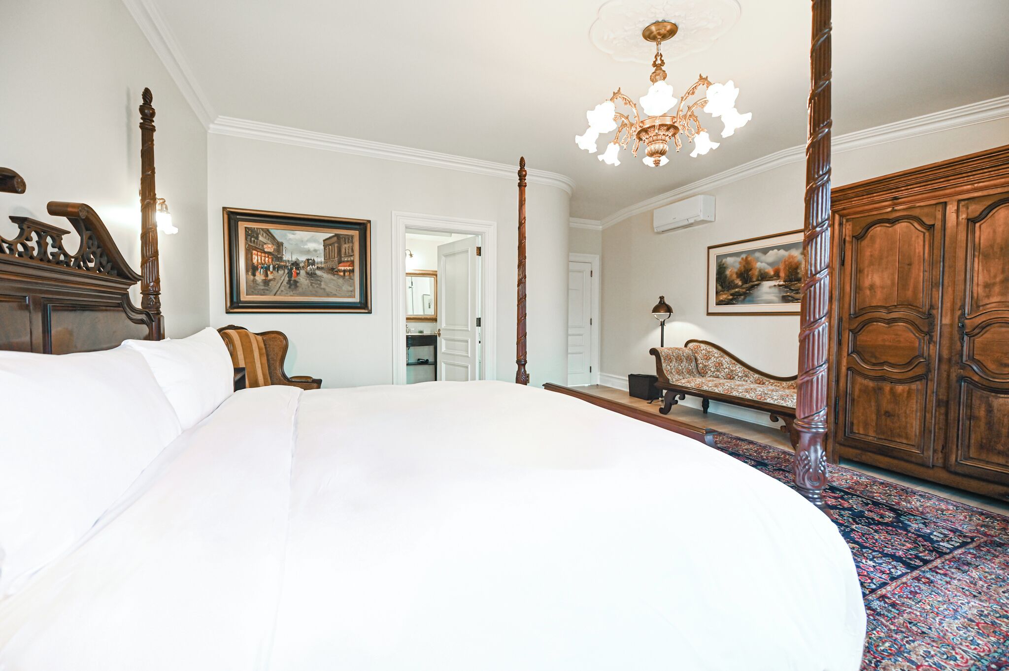 A large bed with a wooden headboard, a chandelier, a wooden wardrobe, and a painting on the wall.