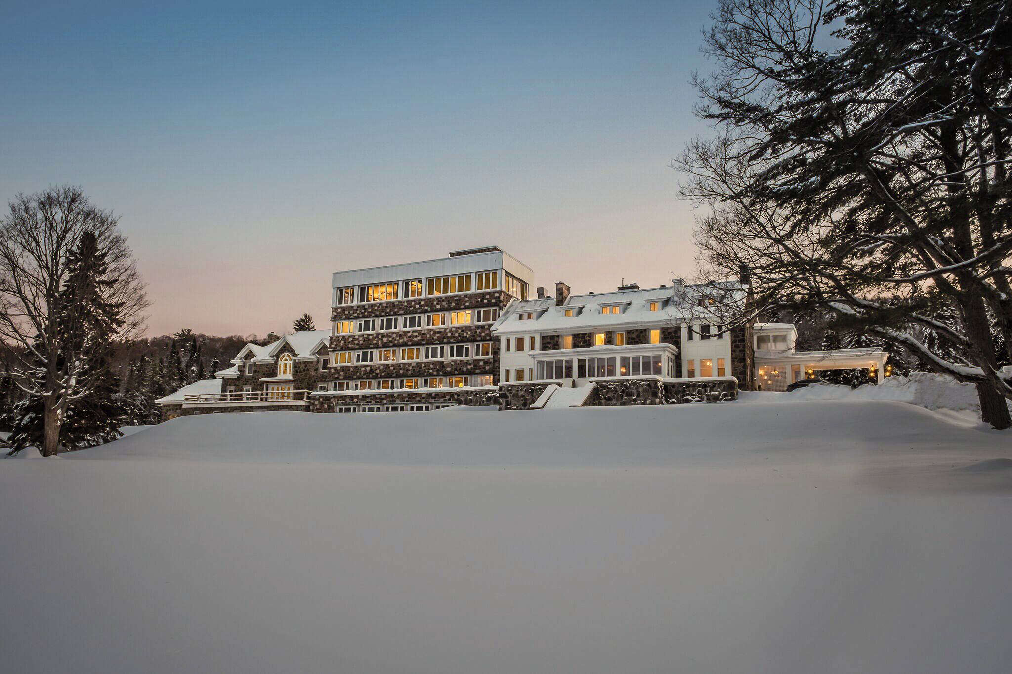 A large, multi-story building with a stone facade and multiple windows, surrounded by snow-covered landscape and trees.
