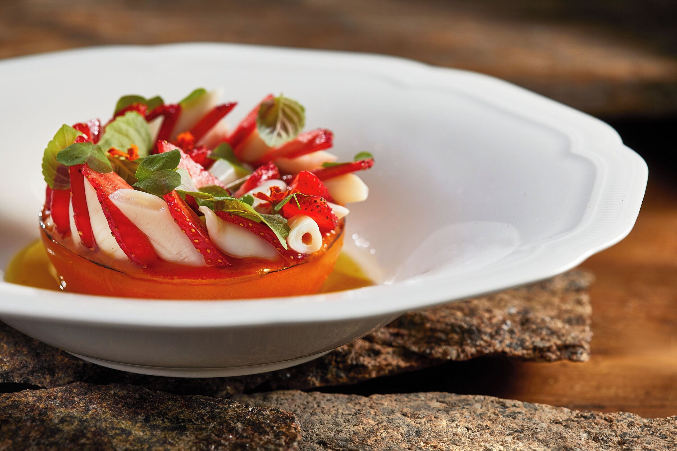Un plato blanco con un guiso a base de tomate, adornado con hierbas y rodajas de fruta roja, sobre una superficie de madera.