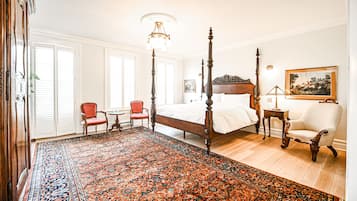 Una cama con dosel y tablero de madera, un sillón rojo, una mesita redonda pequeña y un tapete estampado en una habitación con paredes blancas y suelo de madera.