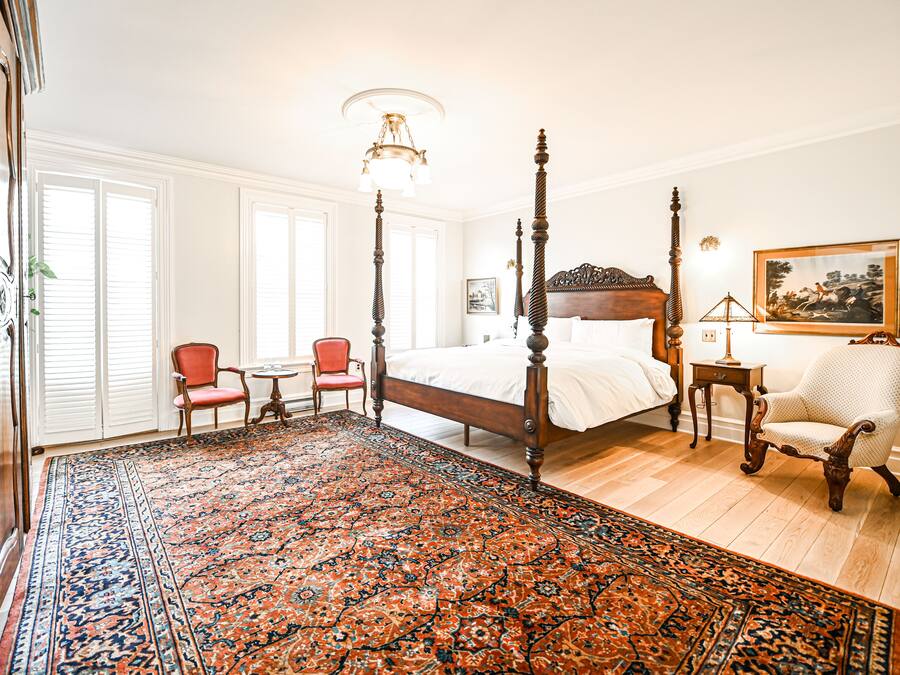 Una cama con dosel y tablero de madera, un sillón rojo, una mesita redonda pequeña y un tapete estampado en una habitación con paredes blancas y suelo de madera.