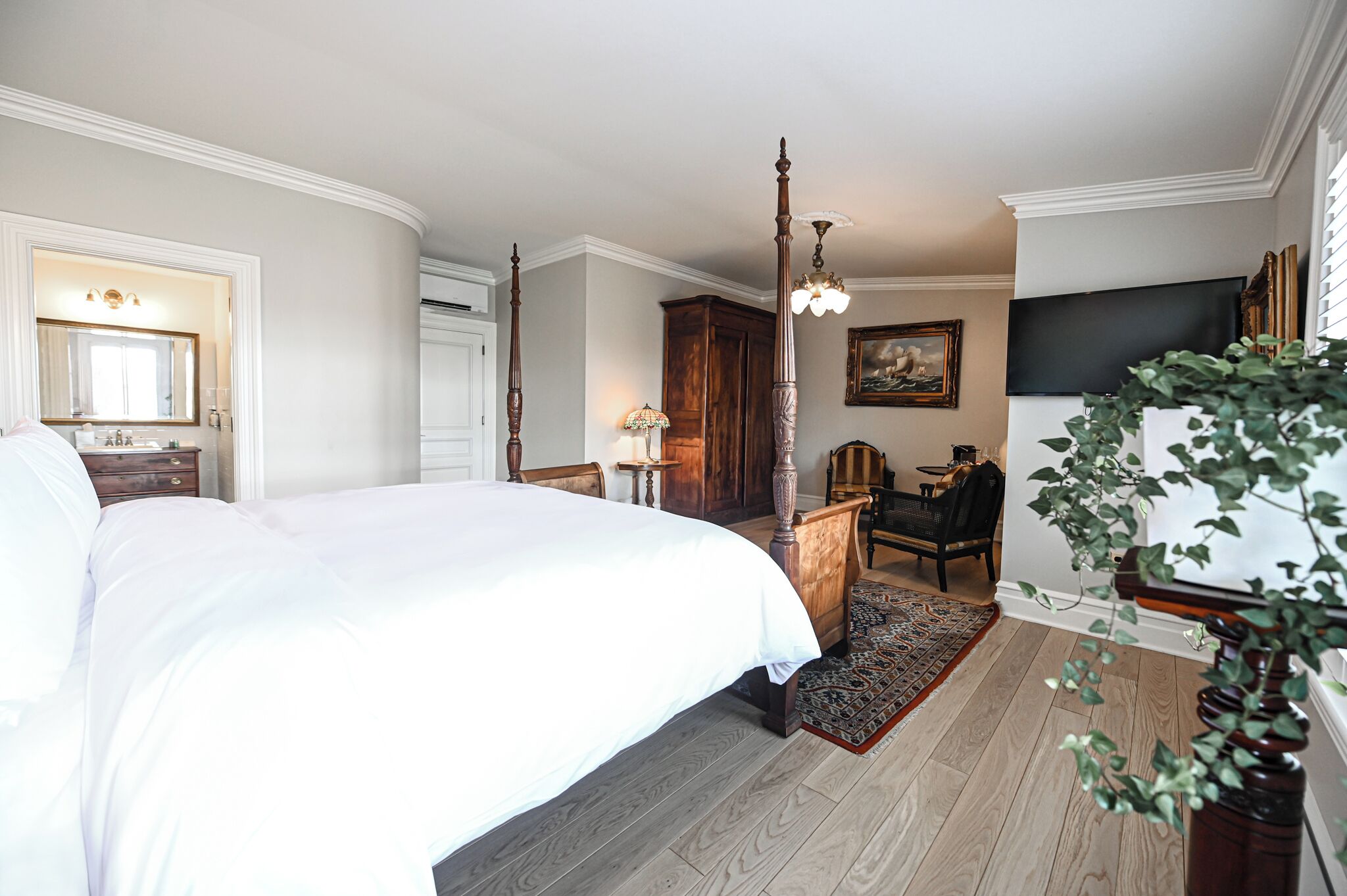 A bedroom with a large bed, a wooden wardrobe, a desk, a chair, a television, and a plant.