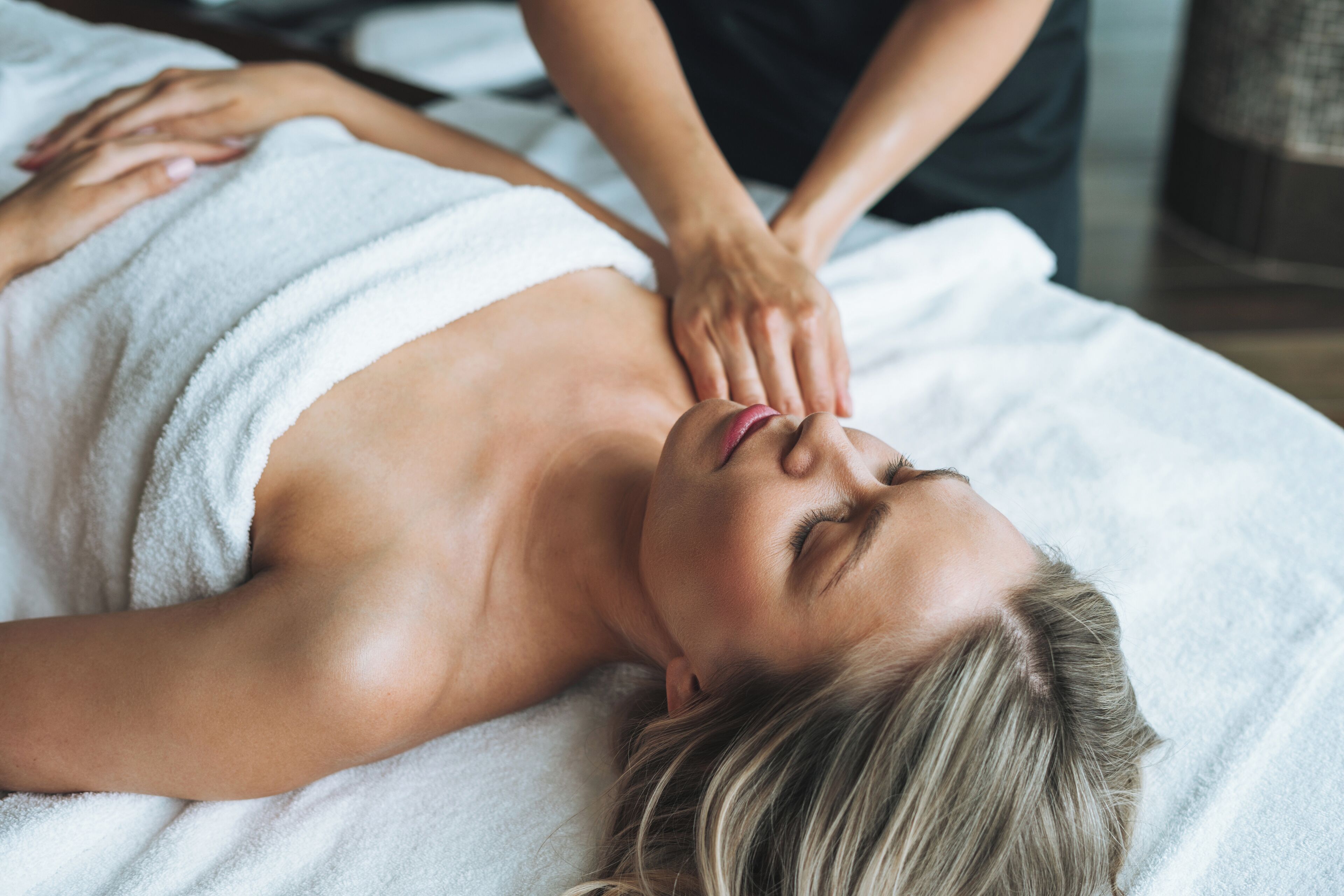 Uma pessoa deitada em uma mesa de massagem com uma toalha enrolada no tronco, recebendo uma massagem nas costas.