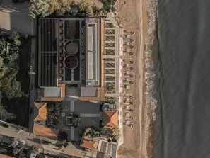 An aerial view of a coastal hotel with a rooftop pool, beachside seating, and a parking area.
