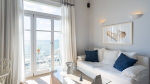 A modern living room with a glass coffee table, white sofa with blue cushions, a framed abstract painting, and a view of the sea through large glass doors. - Dexamenes Seaside Hotel (Ilida)