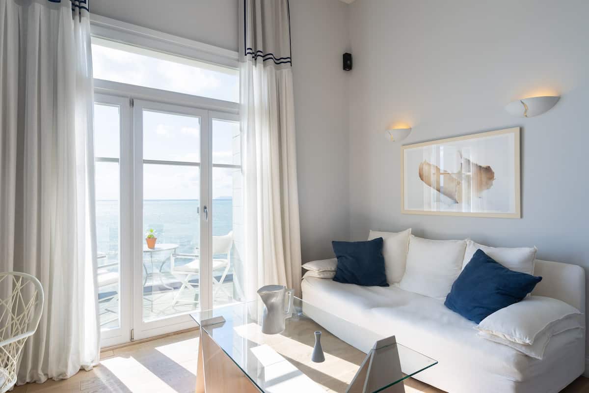 A modern living room with a glass coffee table, white sofa with blue cushions, a framed abstract painting, and a view of the sea through large glass doors.