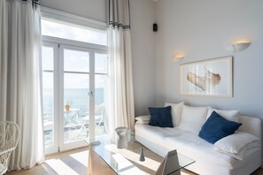 A modern living room with a glass coffee table, white sofa with blue cushions, a framed abstract painting, and a view of the sea through large glass doors. - Dexamenes Seaside Hotel (Ilida)