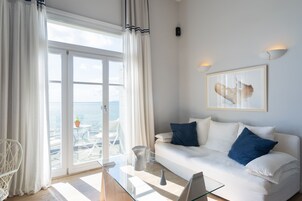 Una sala de estar moderna con una mesa de centro de vidrio, sofá blanco con cojines azules, un cuadro abstracto enmarcado y vista al mar a través de amplios ventanales.