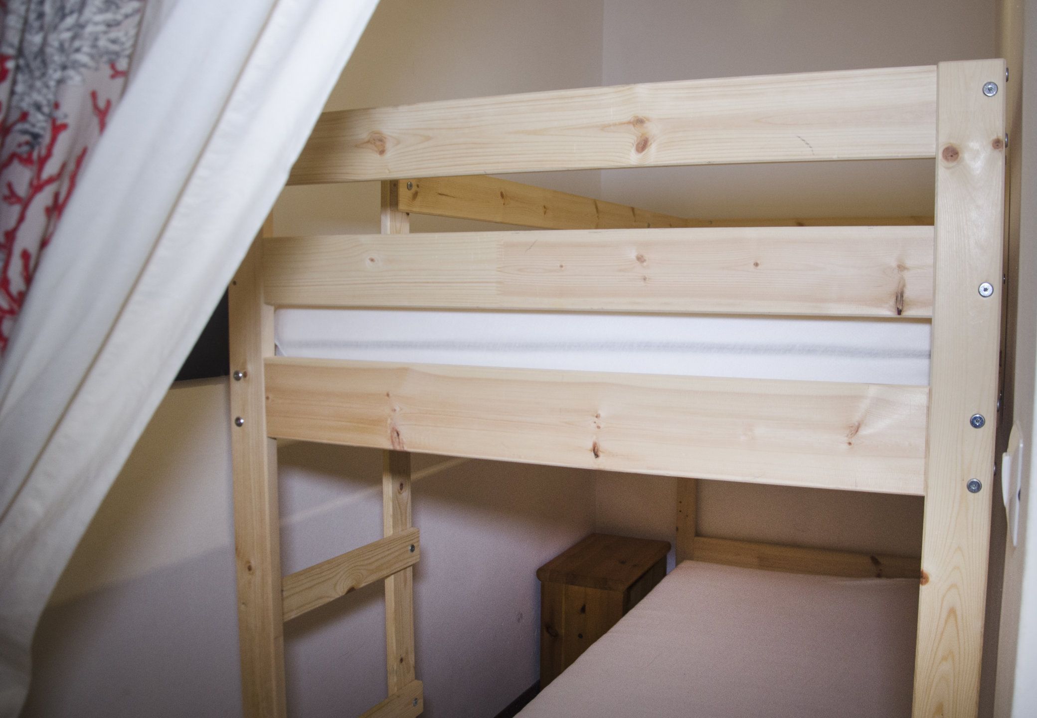A wooden bunk bed with a ladder, a small wooden chest, and a white curtain.