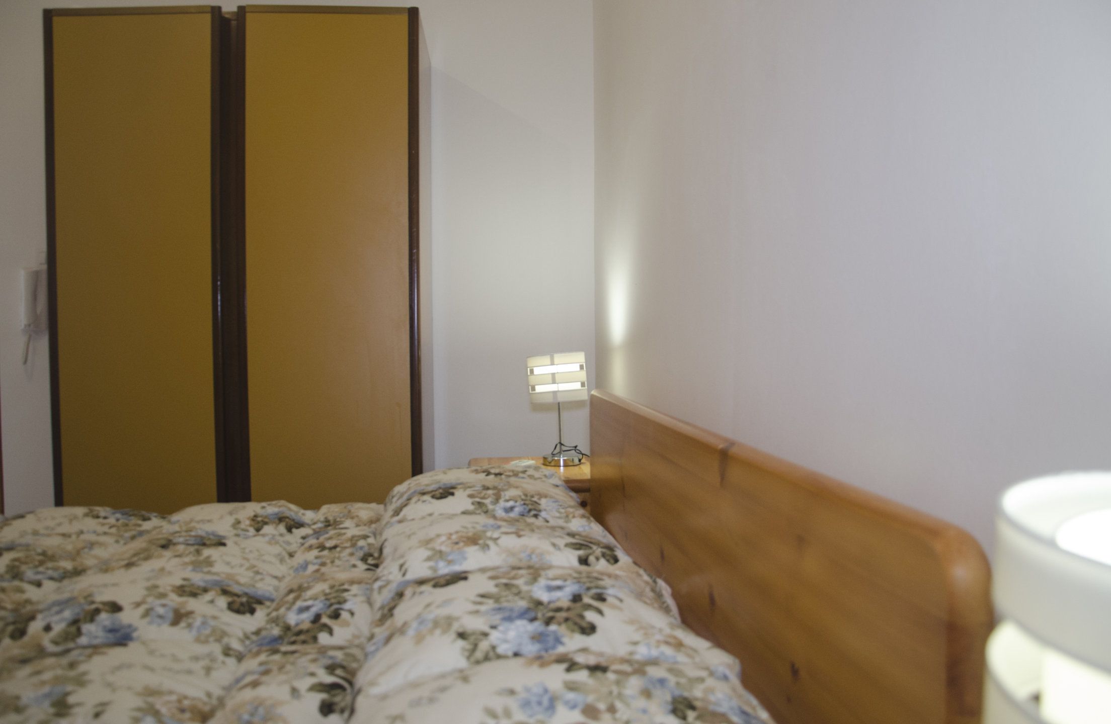 A bedroom with a wooden headboard, a bed with floral bedding, a nightstand with a lamp, and a wardrobe with a mirror.