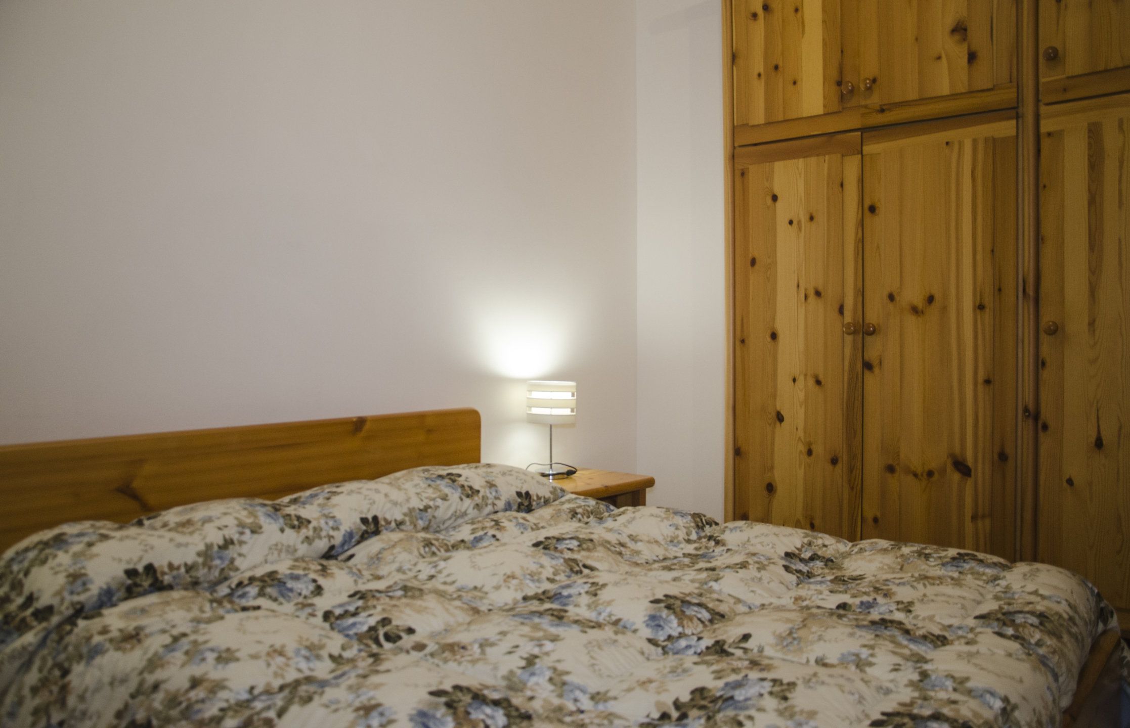 Une chambre à coucher avec une tête de lit en bois, un lit recouvert d’une couvre-lit à motifs floraux, une lampe de chevet et une armoire en bois.