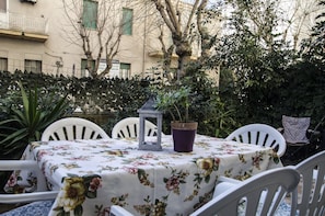 Un patio al aire libre con una mesa redonda cubierta con un mantel estampado de flores, rodeada de sillas blancas.