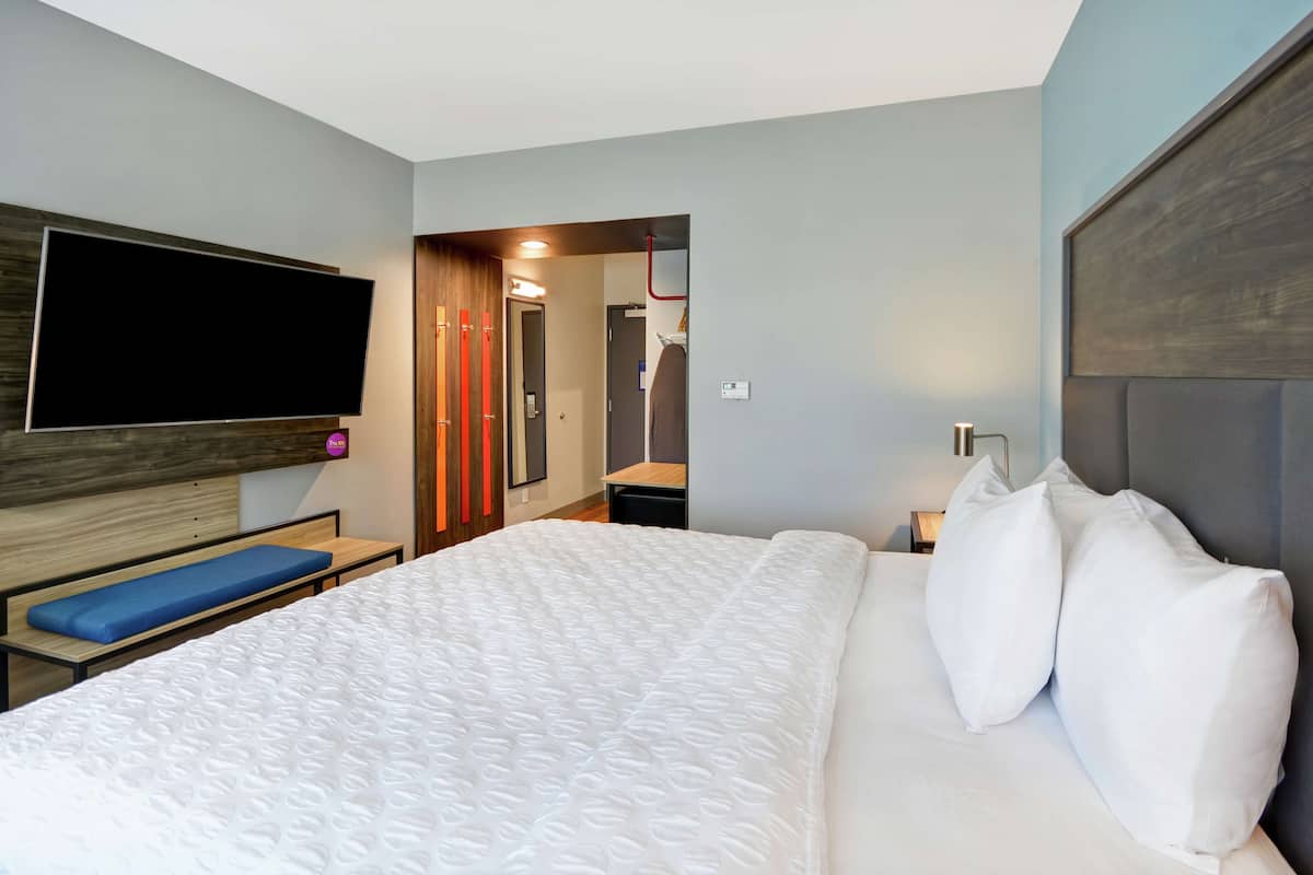 Une chambre d’hôtel moderne dotée d’un grand lit, d’un téléviseur à écran plat fixé au mur, d’une banquette au pied du lit et d’un dressing.