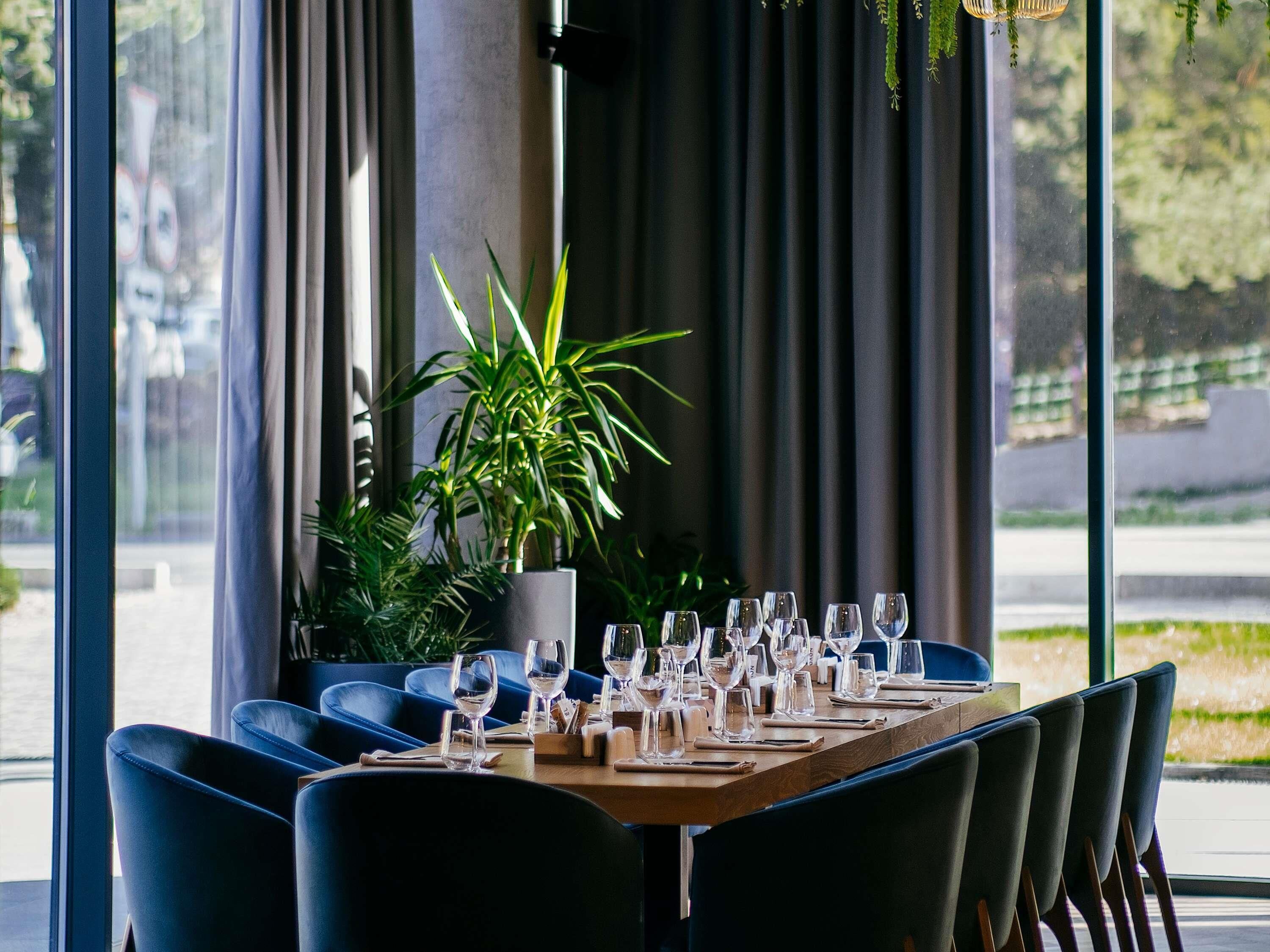 Un restaurante con una larga mesa de madera dispuesta para comer, rodeado de sillas azules y una gran planta en maceta en el centro.