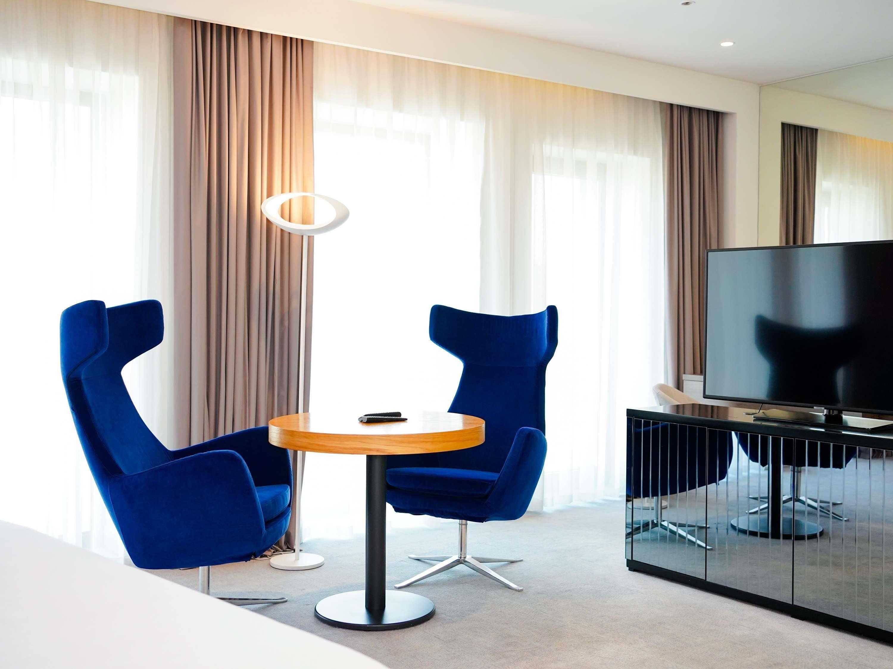 Een moderne hotelkamer met een ronde tafel, twee blauwe stoelen, een televisie en een glazen scheidingswand.
