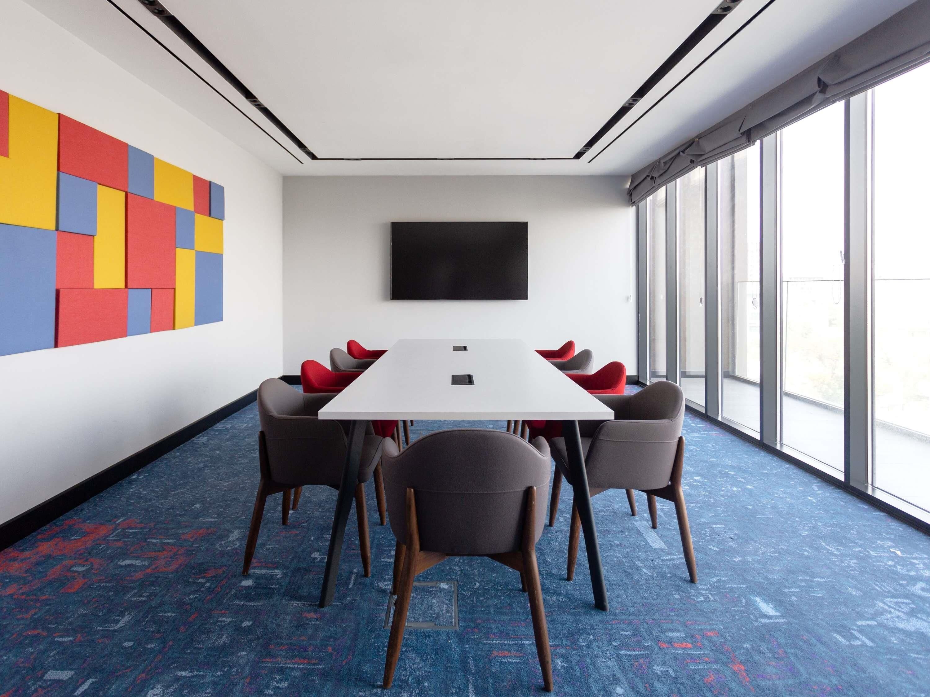 Un salón de conferencias con una mesa blanca, seis sillas, un ventanal grande y una obra de arte colorida en la pared.