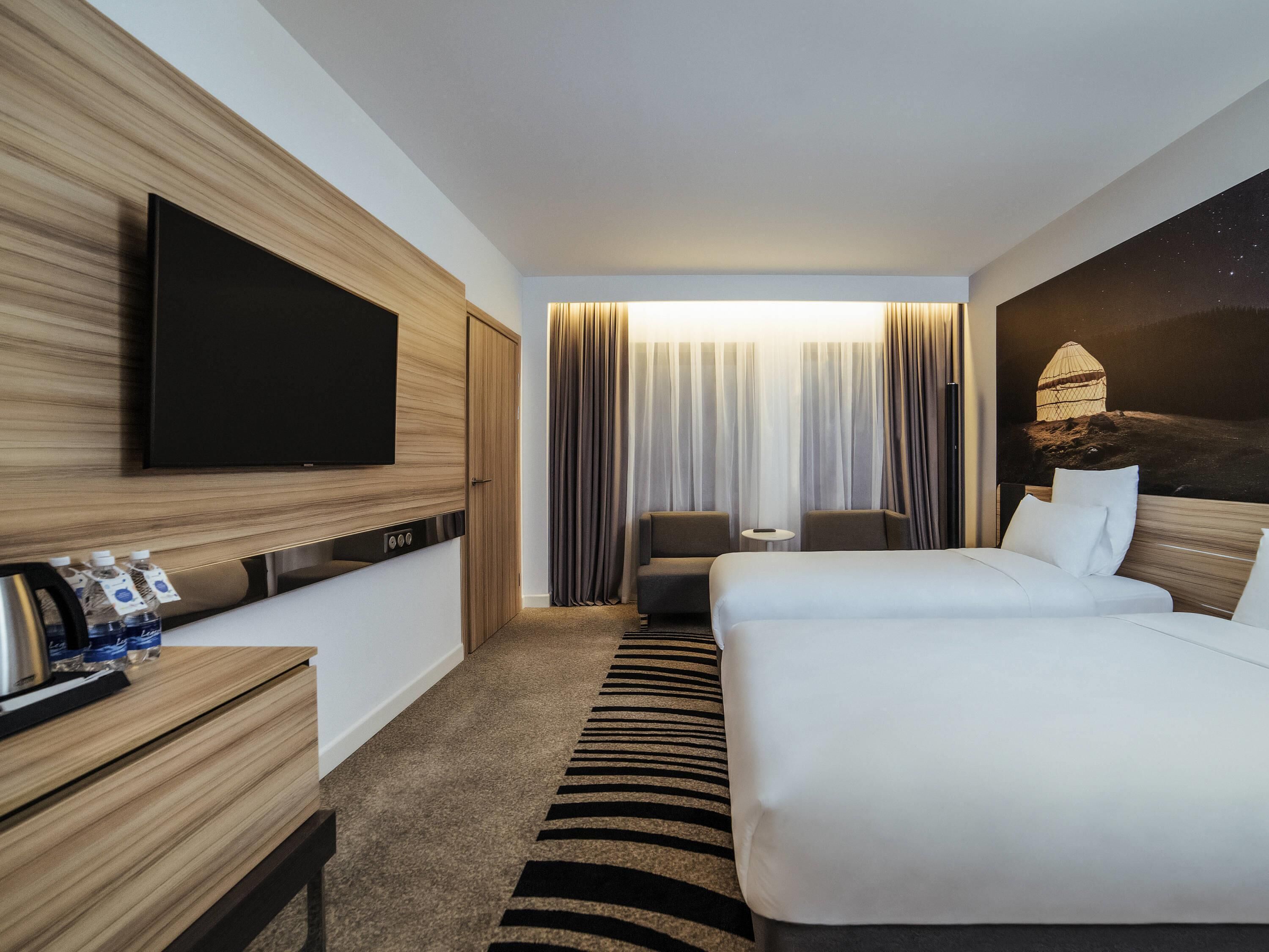 Ein Hotelzimmer mit einem Bett, einem Fernseher, einem Schreibtisch mit einer Kaffeemaschine, einem Sessel und einem Fenster mit Vorhängen.