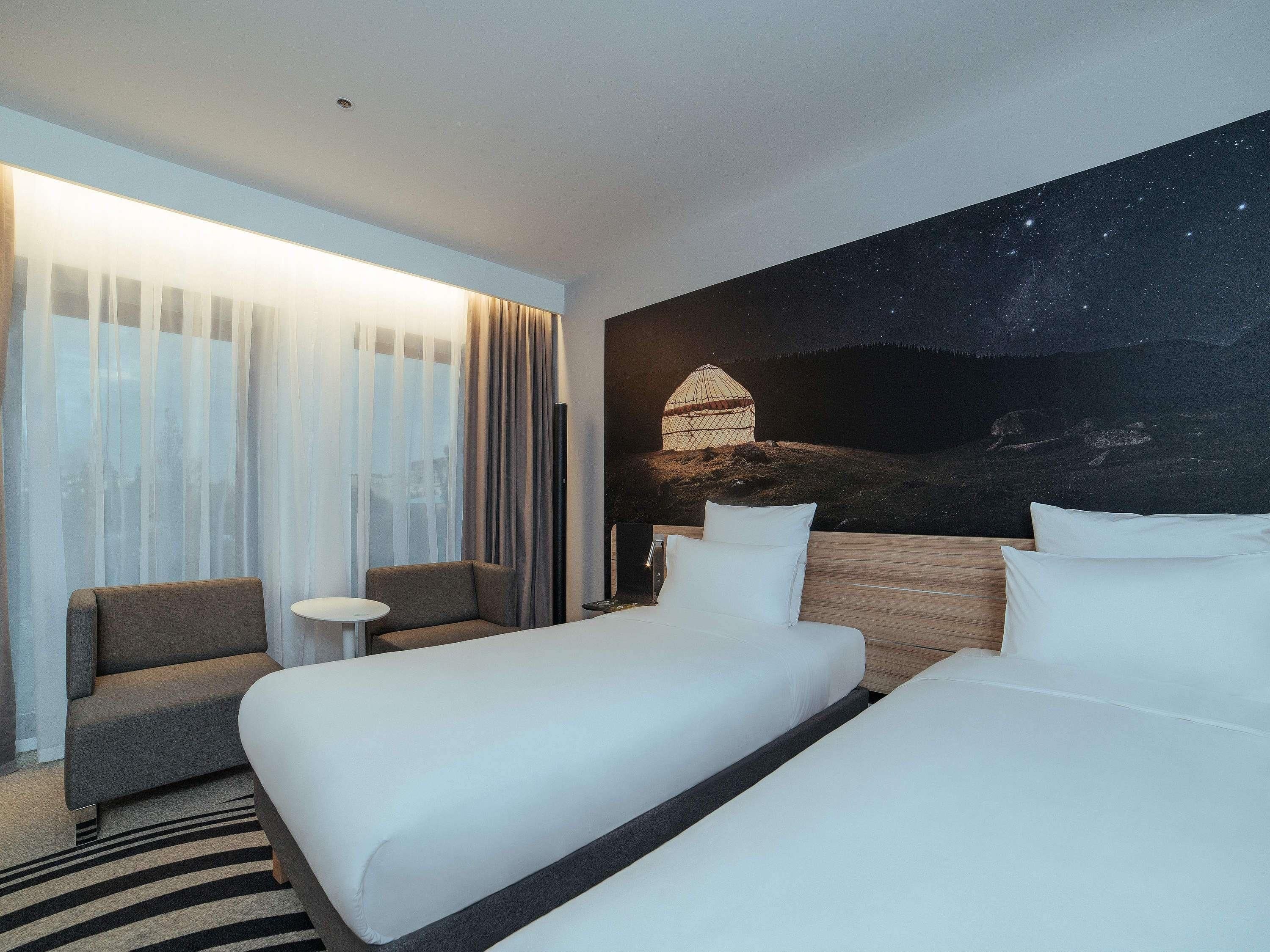 A hotel room with two beds, a sofa, a small table, and a mural of a night sky with a yurt.