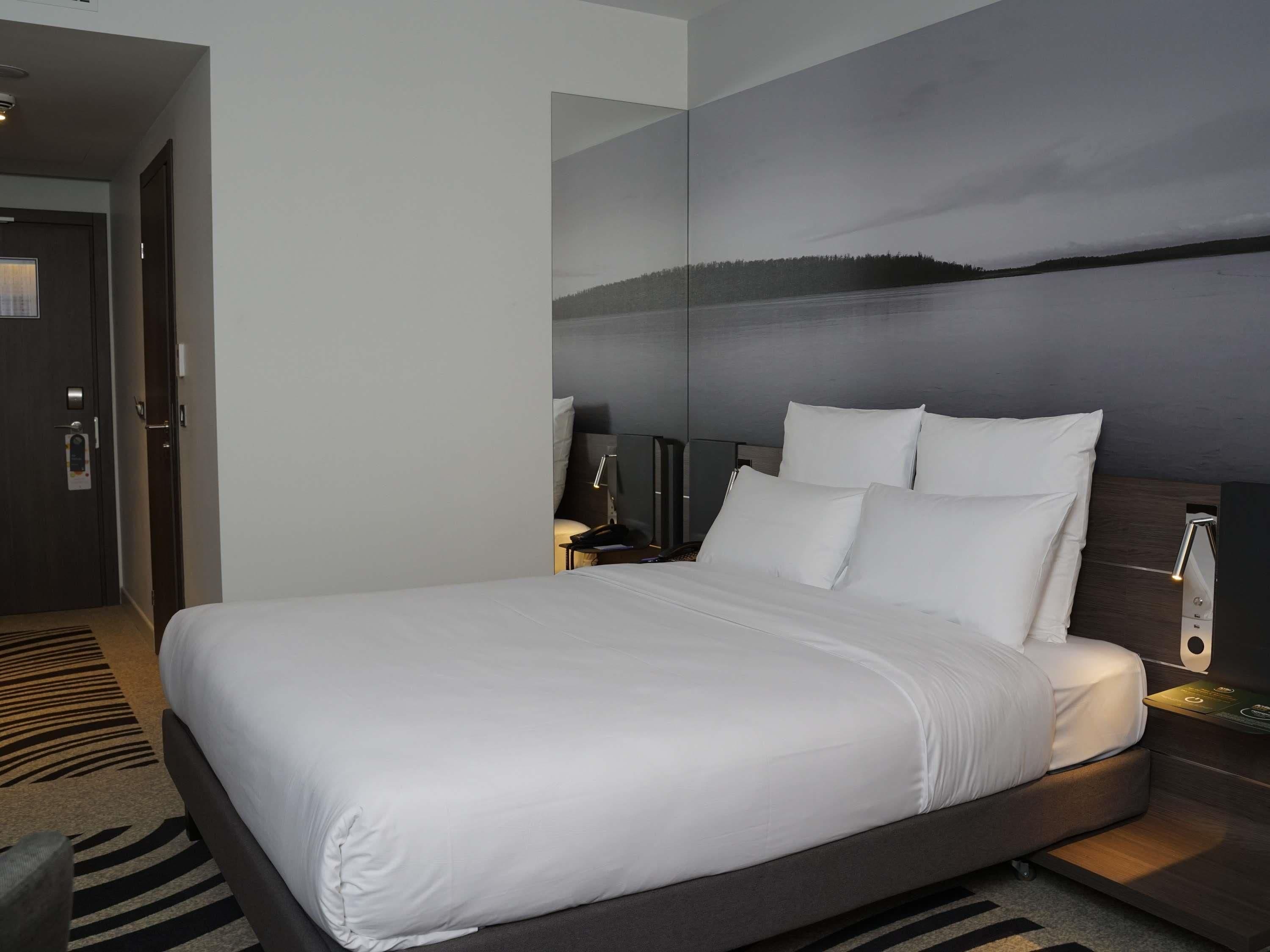 A modern hotel room with a large bed, a nightstand, a lamp, and a view of a lake through a large window.