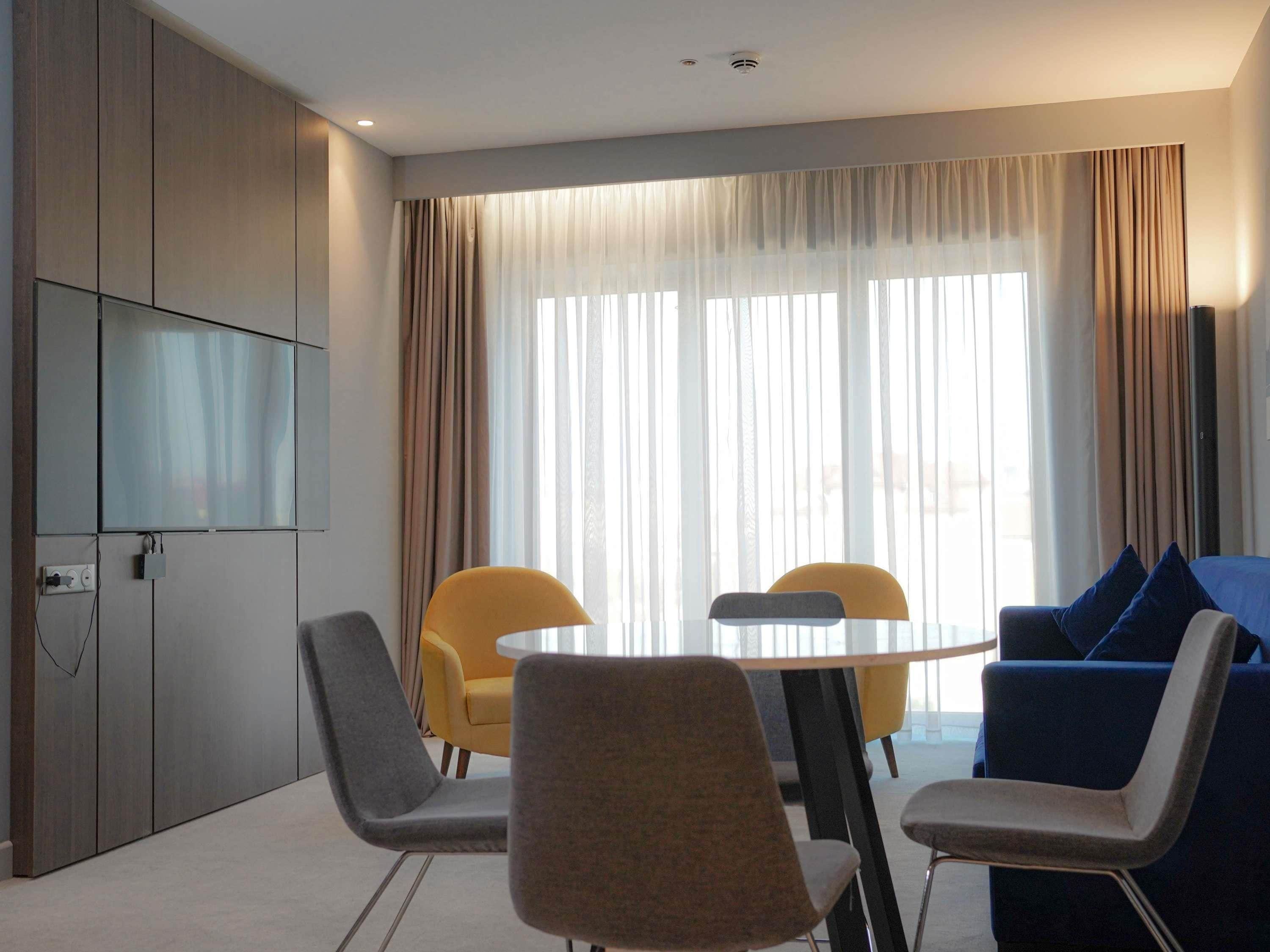 A modern hotel room with a dining area, a flat-screen TV mounted on the wall, and a large window with curtains.