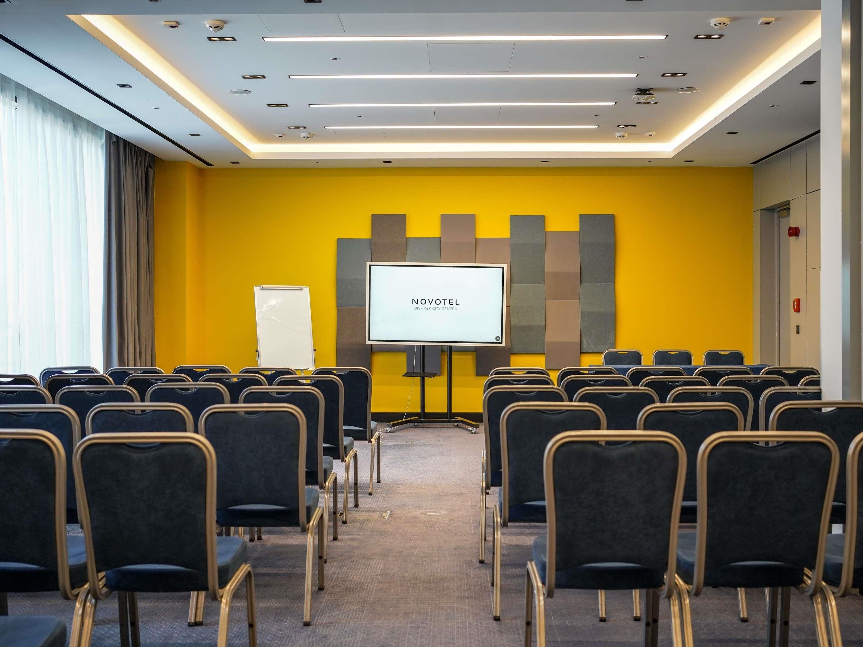 Une salle de conférence avec des rangées de chaises, un tableau blanc et un écran affichant le mot « NOVOTEL ».