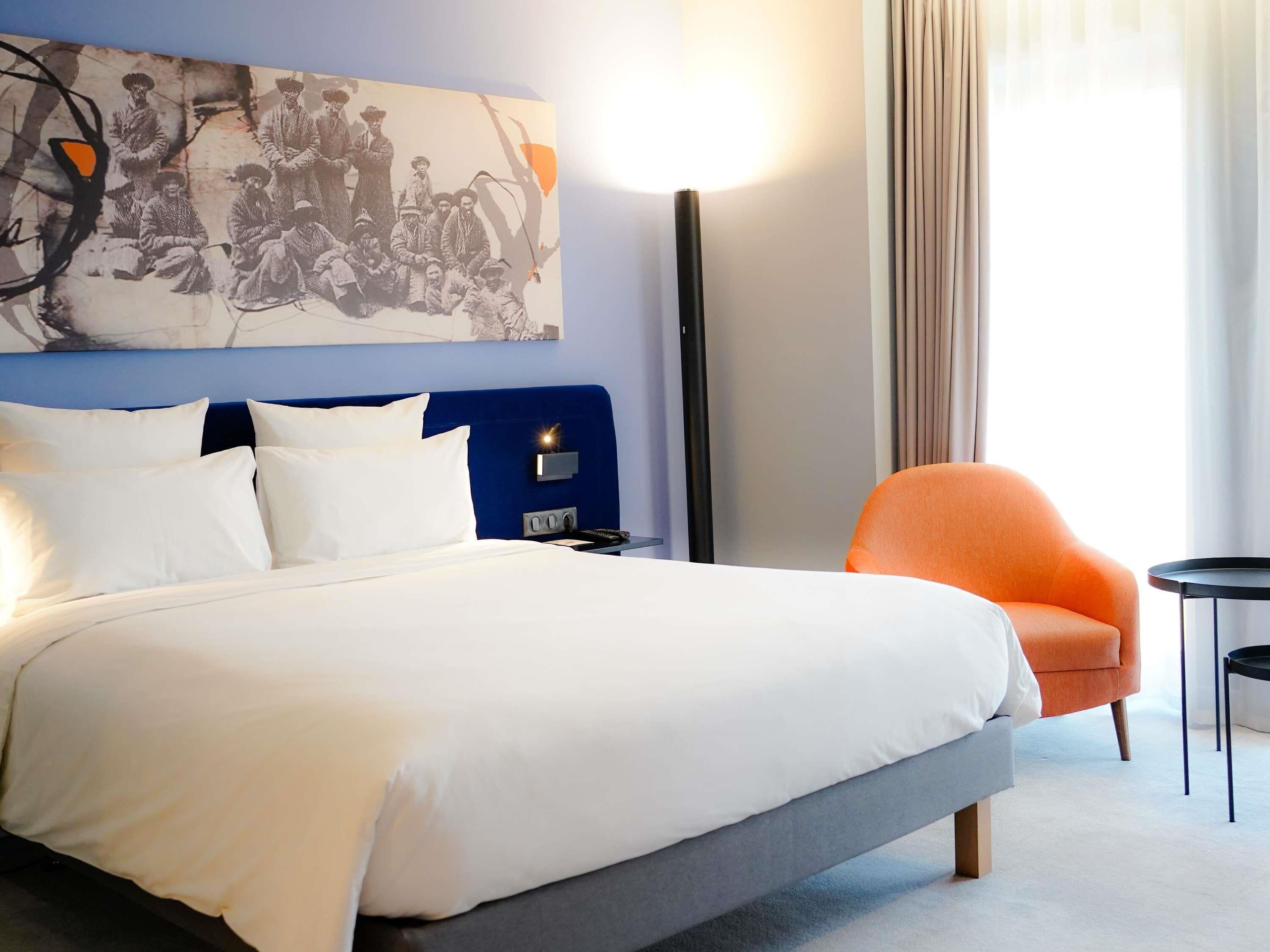 Une chambre d’hôtel moderne avec un grand lit, une tête de lit bleue, une table de chevet, une chaise orange et un lampadaire.