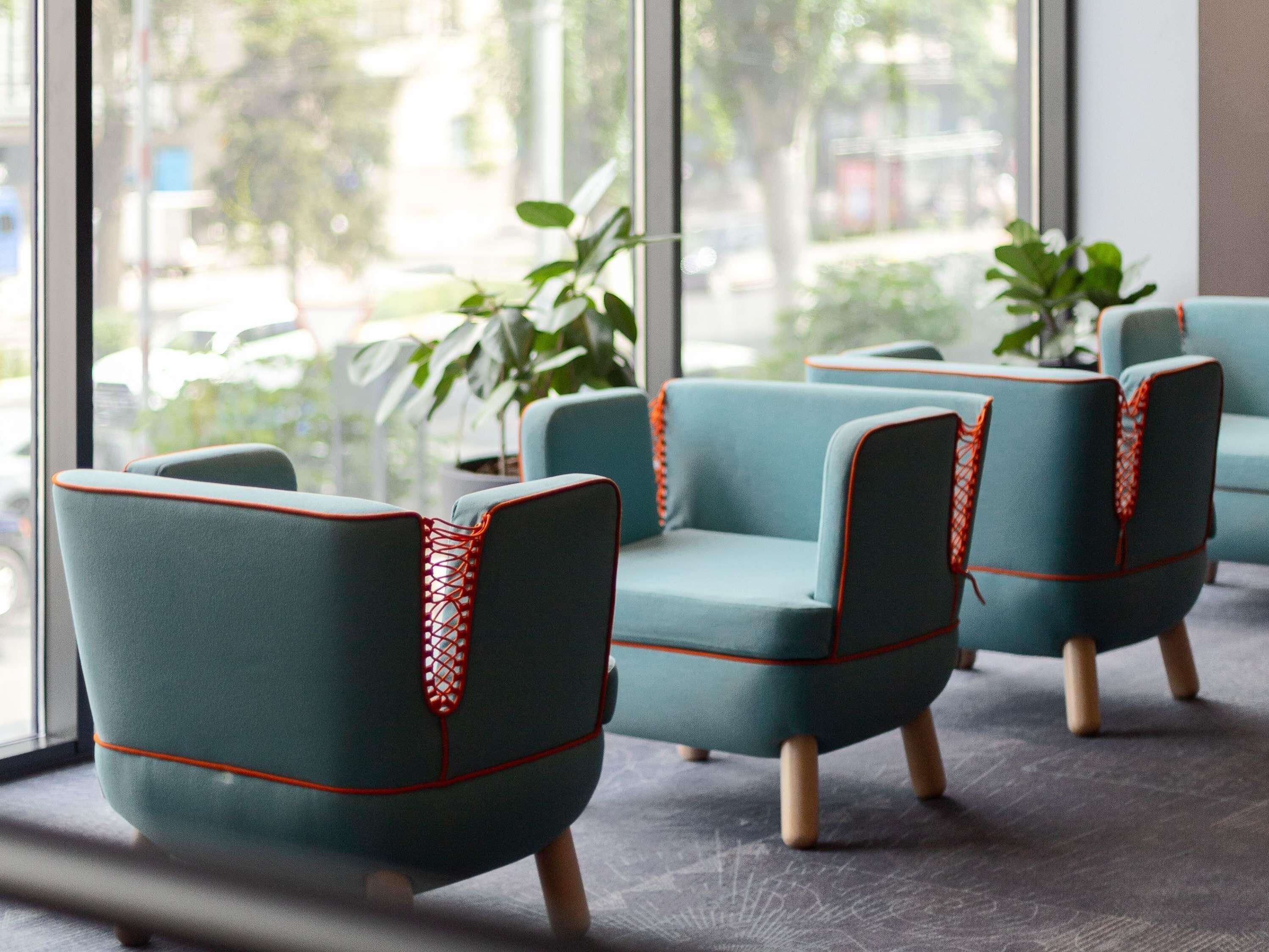 A row of teal chairs with wooden legs and red trim, positioned in front of a large window with a view of greenery and buildings.
