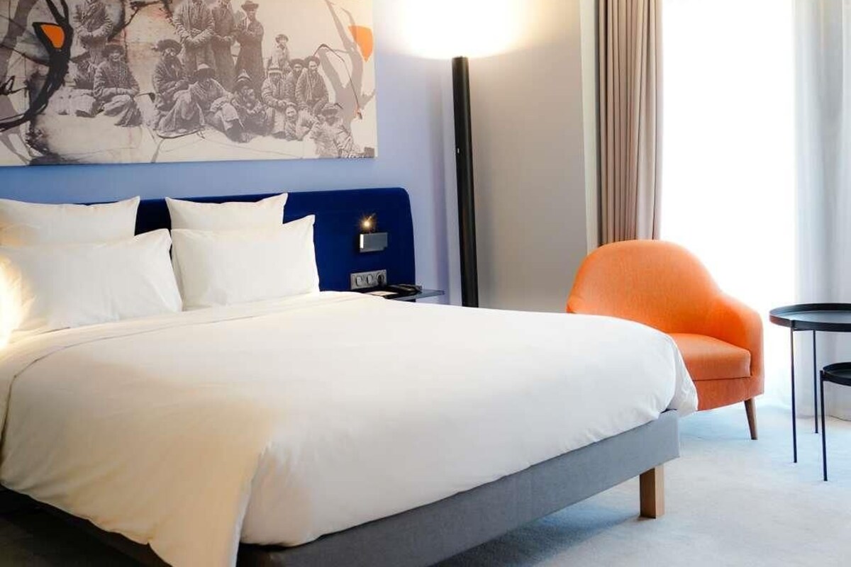 A hotel room with a bed, a blue headboard, an orange armchair, a small table, and a wall-mounted artwork.