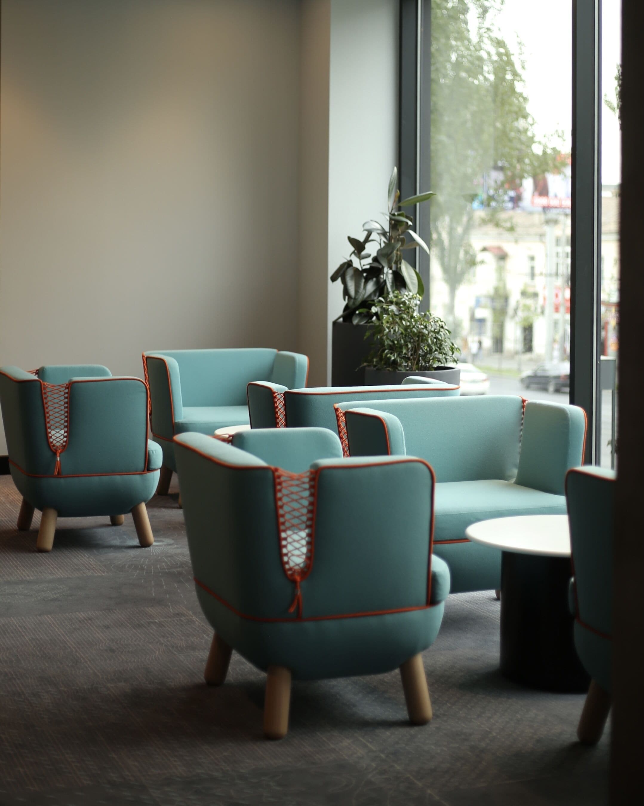 A modern interior with teal armchairs and a sofa, a round table, and a large window with a view of the city.
