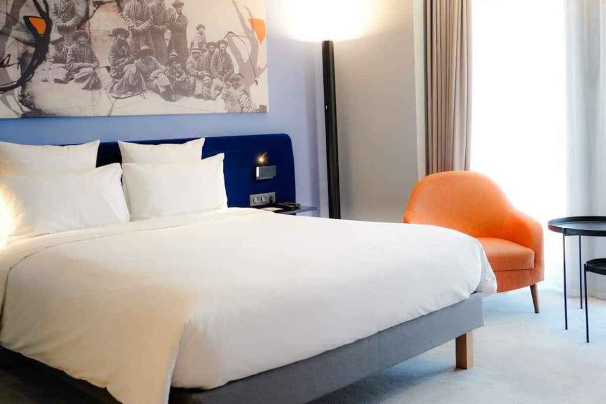 A hotel room with a bed, a blue headboard, an orange armchair, a small table, and a wall-mounted artwork.