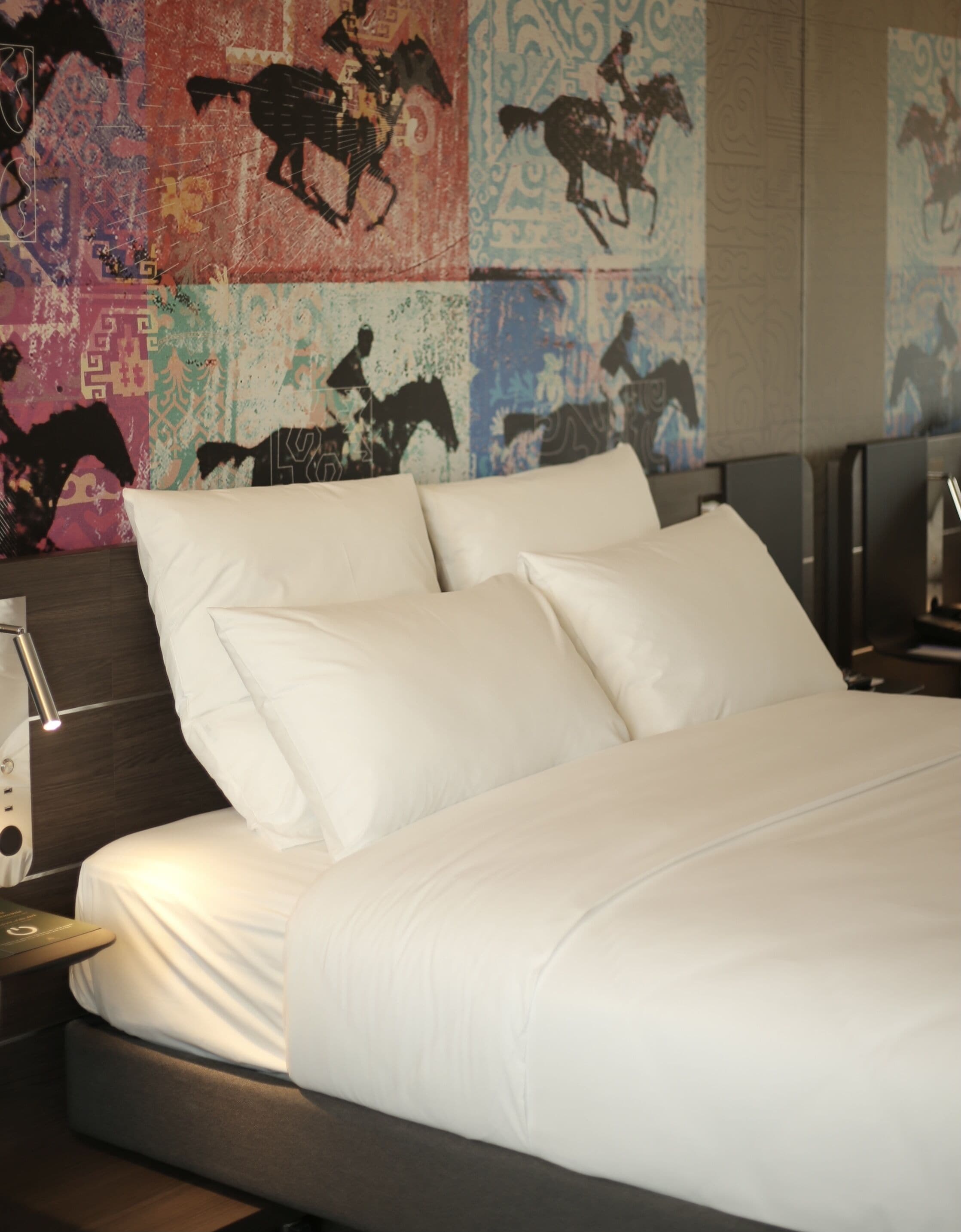 A neatly made bed with white linens, a modern headboard, and a wall decorated with horse-themed artwork.