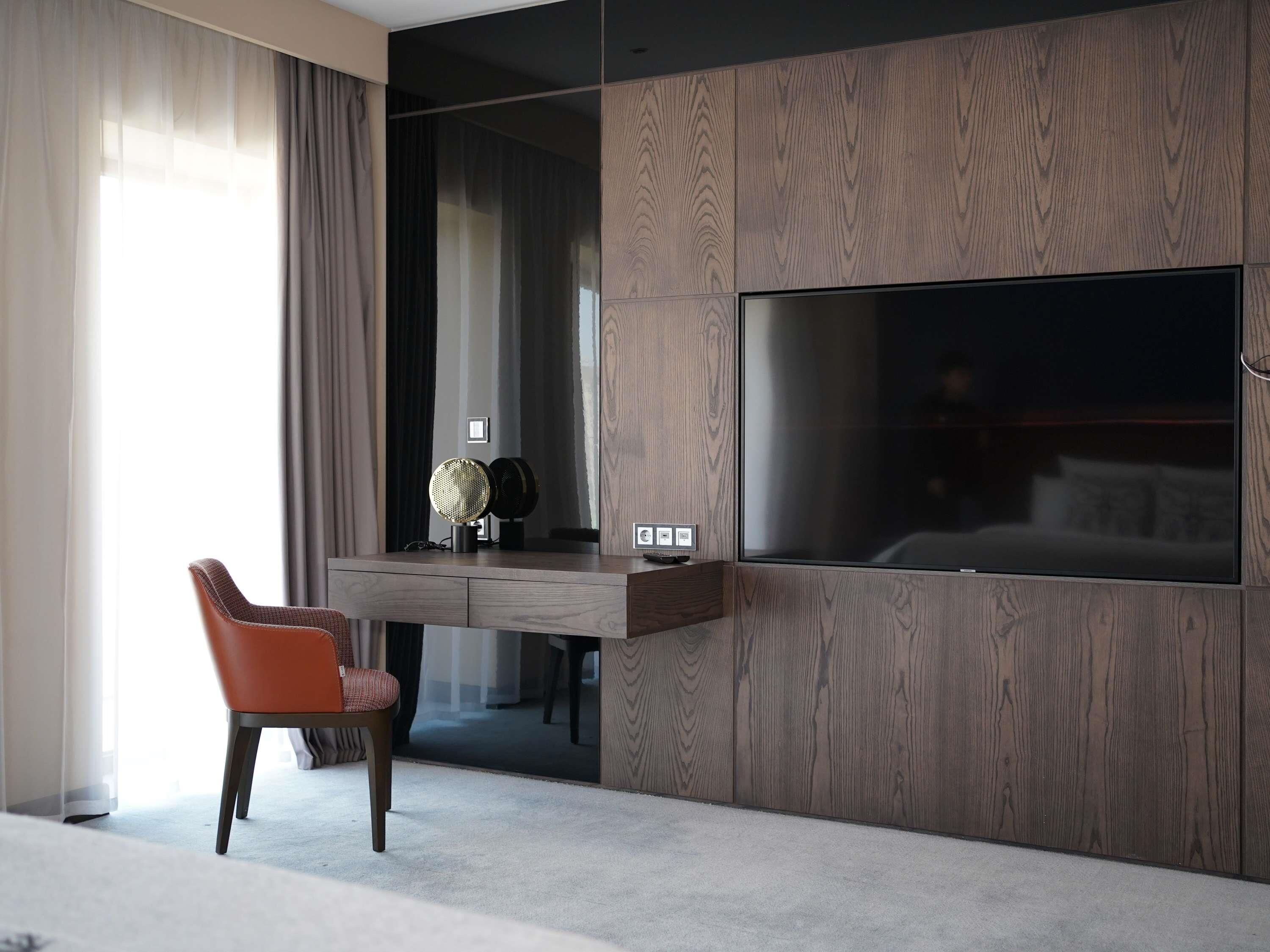 A modern hotel room with a large flat-screen TV mounted on a wooden panel wall, a comfortable orange chair, and a small table with decorative items.