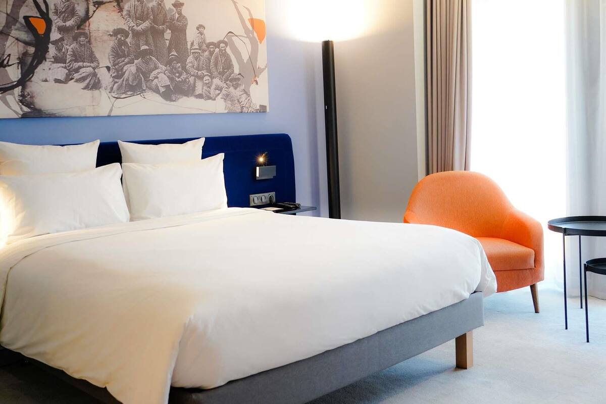 A hotel room with a bed, a blue headboard, a painting, a standing lamp, an orange armchair, and a small table.
