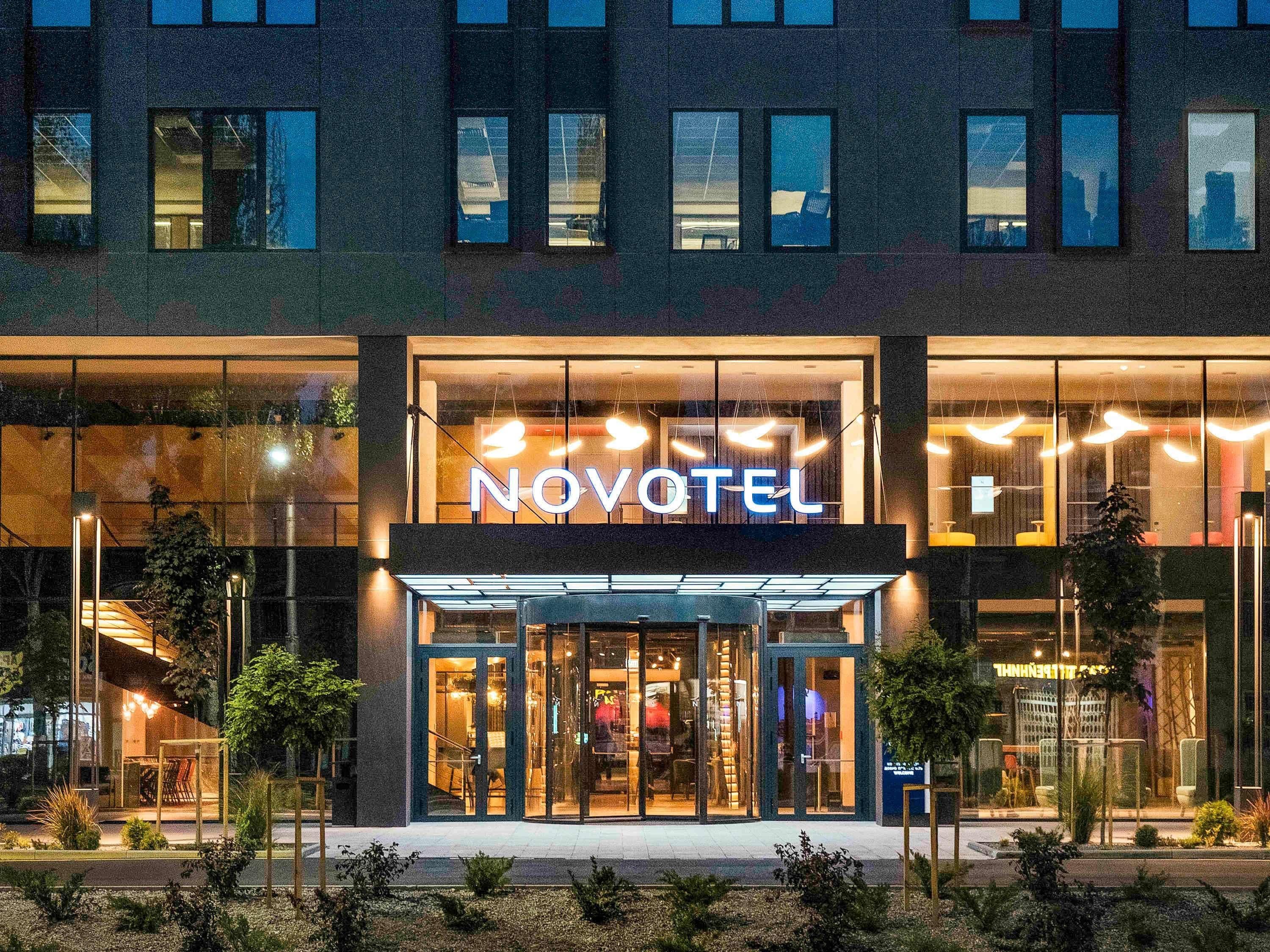 A modern hotel with a prominent 'NOVOTEL' sign, large glass windows, and a well-lit entrance.